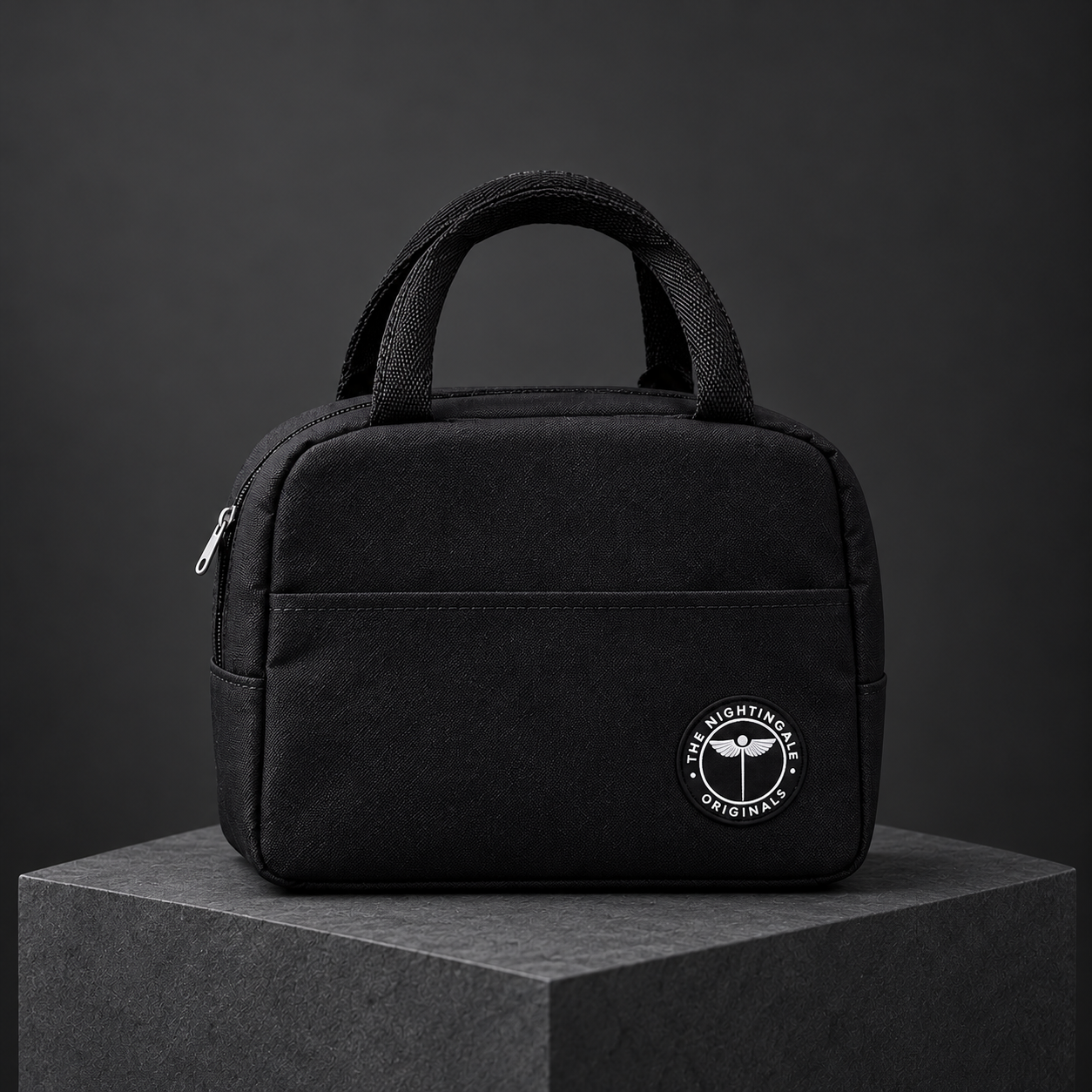 Three-quarter view of a black Famish’d lunch tote with a front pocket on a dark stone plinth.