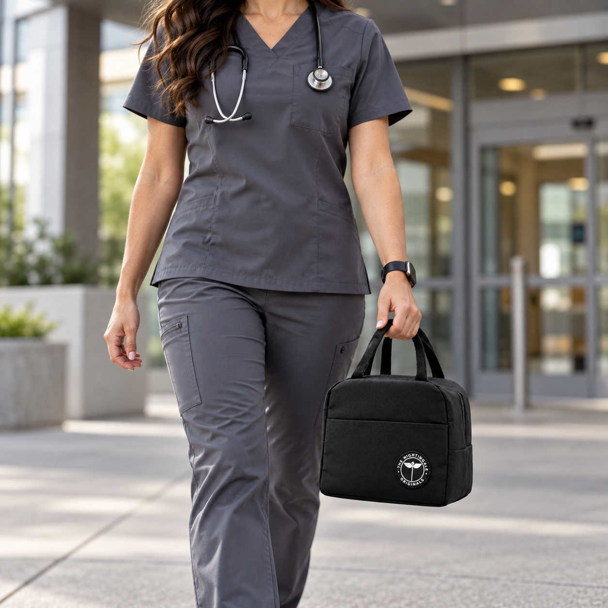 Person wearing gray scrubs carrying a black Famish’d lunch tote outside a modern building.