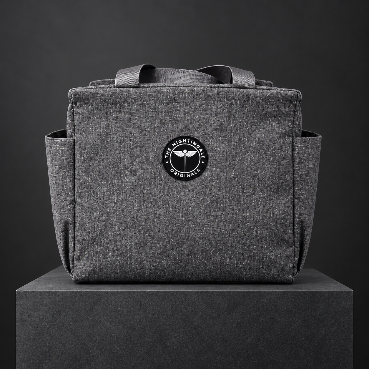 Gray Famish’d 15L lunch bag with handles, a front pocket and round NTG emblem on a dark plinth.