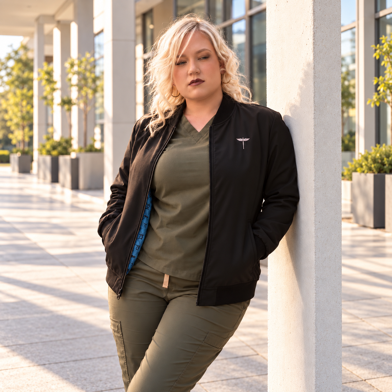 Model wearing a black NTG healthcare bomber jacket over olive scrubs beside a stone column.