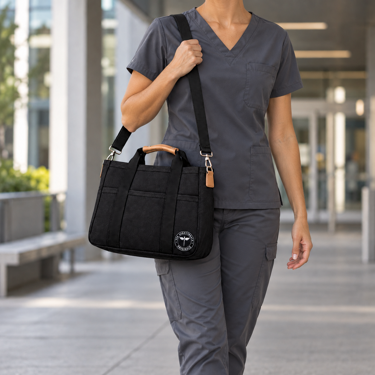 Person in gray scrubs carrying a black Satch’l clinical tote by its tan handles outside a building.