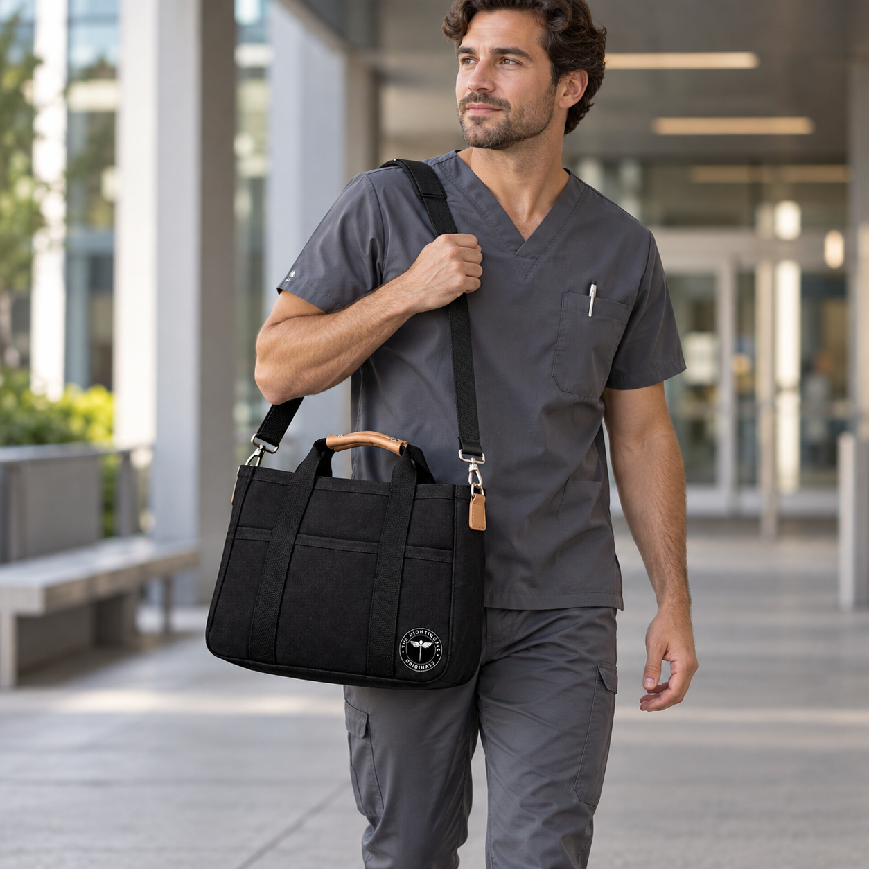 Model in gray scrubs carrying a black Satch’l clinical tote over one shoulder outside a building.