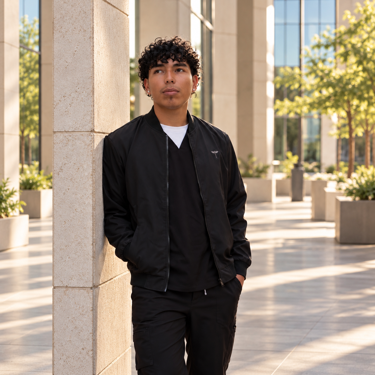 Model wearing a black NTG healthcare bomber jacket over black scrubs beside a stone column.
