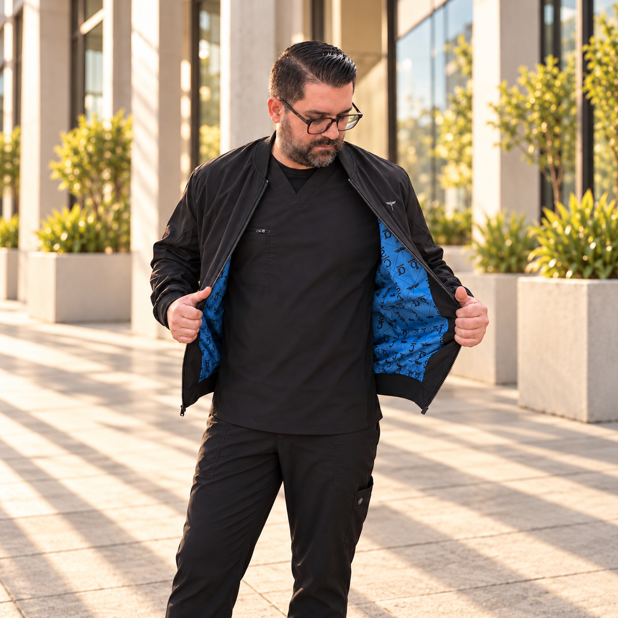 Model in black scrubs holding open a Venous bomber jacket with blue printed lining beside stone columns.